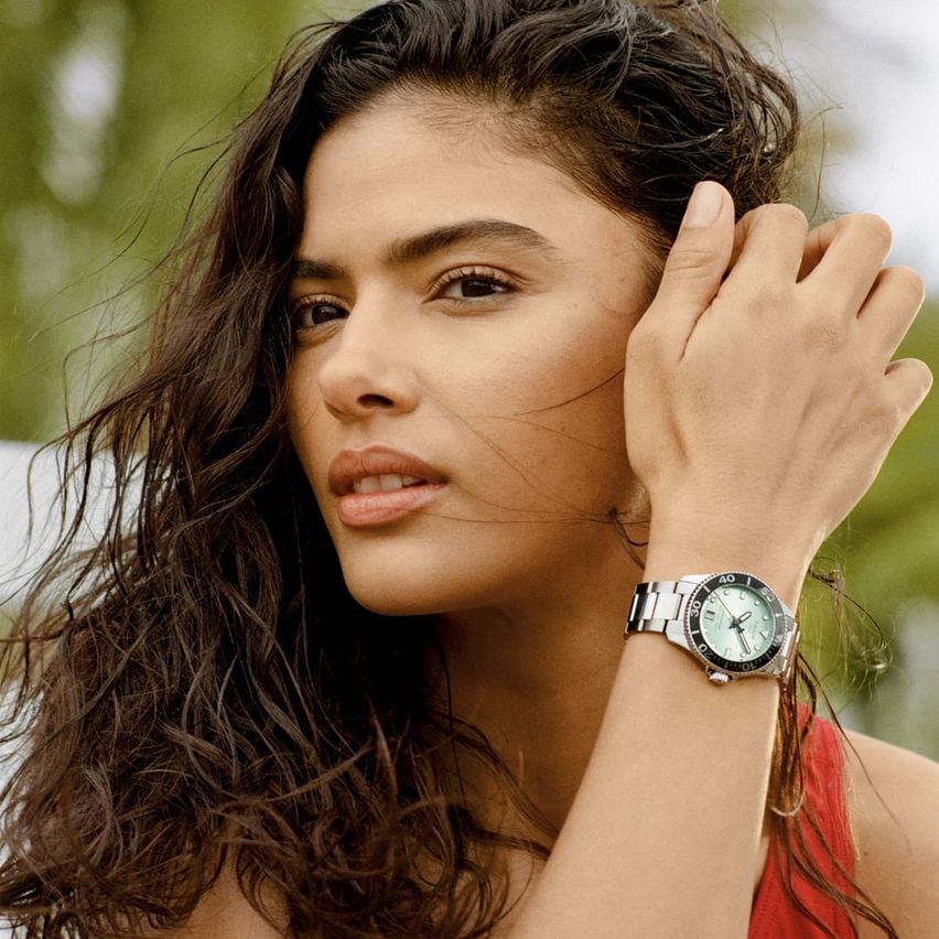 Eine Frau mit lockigem Haar trägt ein rotes Oberteil und eine silberne Uhr am Handgelenk. Sie berührt ihr Haar mit der Hand.