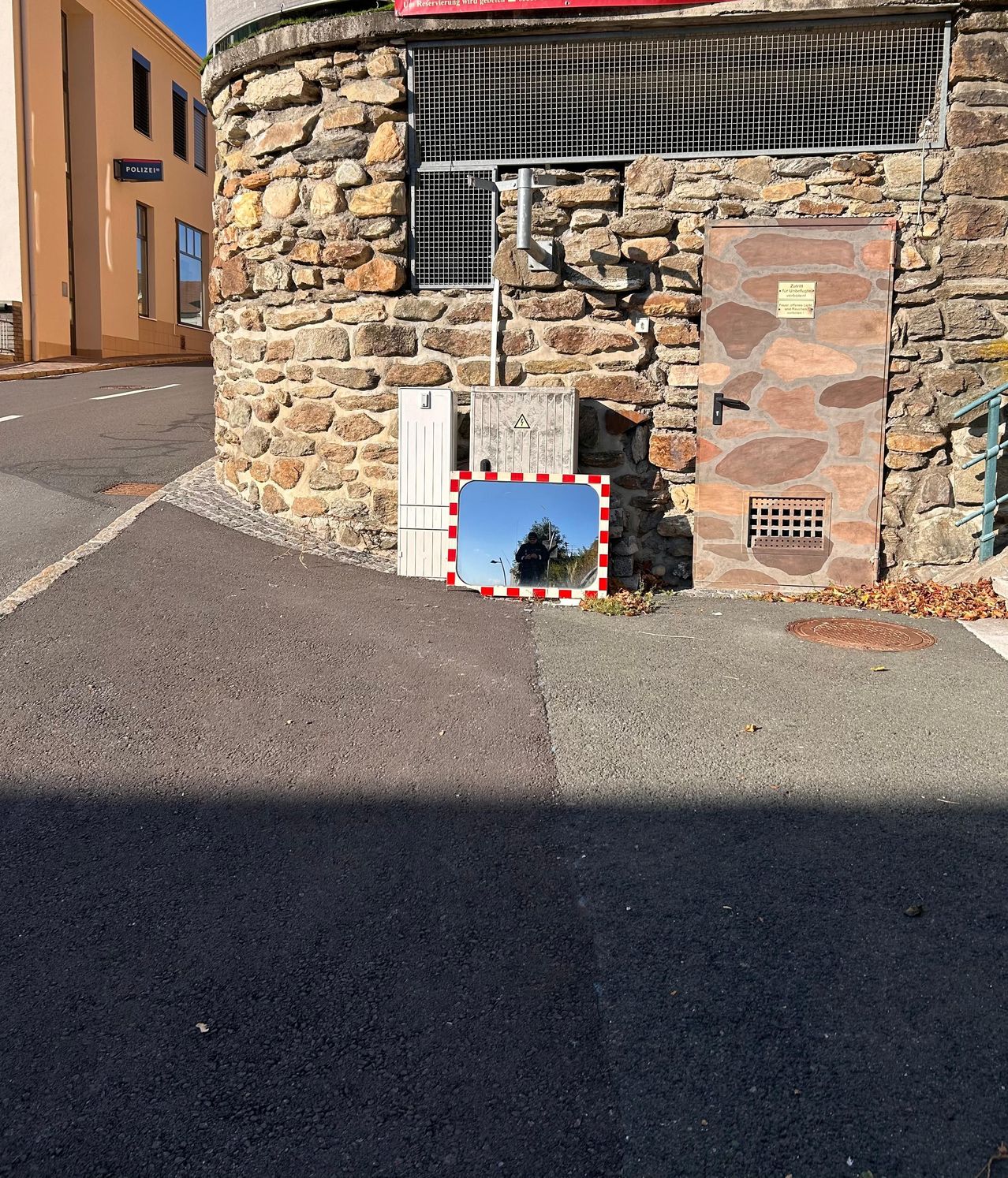 Eine Straßenecke mit einem reflektierenden Spiegel und einer Steinmauer. Es gibt eine Tür und ein Schild an der Wand. Ein Gebäude befindet sich auf der linken Seite.