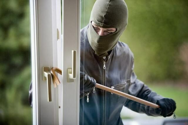 Eine Person mit grüner Maske, Handschuhen und einem schwarzen Mantel versucht, ein Haus mit einem Holzstock zu betreten.