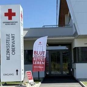 Der Eingang zu einem Gebäude hat zwei große weiße Schilder. Ein Schild hat den Text 'Bezirksstelle Oberwart' und einen nach oben weisenden Pfeil. Das andere Schild hat ein rotes Kreuz mit den Worten 'Blut Spenden Retten Leben'. Vor dem Eingang befindet sich eine Fahne mit einem roten Kreuz.