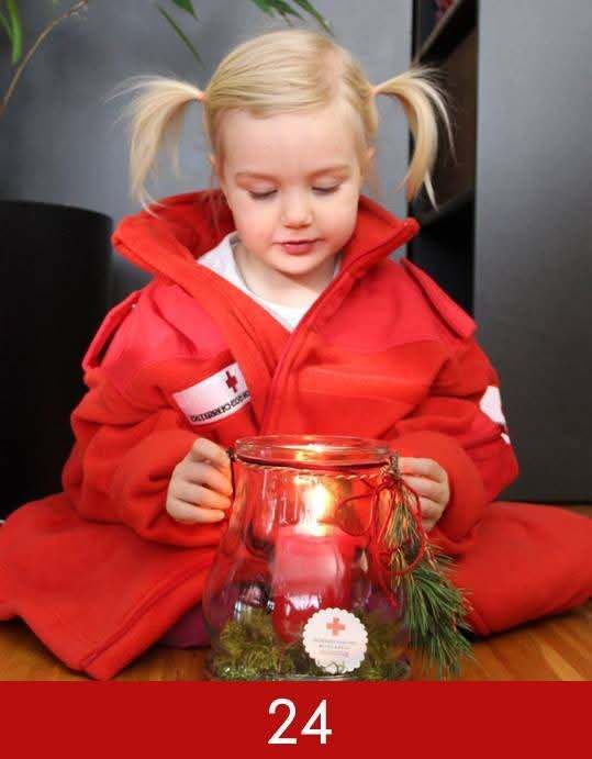 Ein kleines Kind, das einen roten Mantel mit einem weißen Kreuzsymbol auf der linken Seite trägt, hält eine Kerze in einem Glas mit rotem Deckel. Das Kind sitzt auf dem Boden.