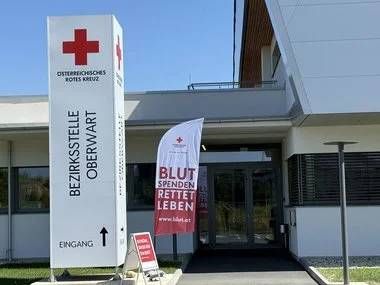 Ein rotes Kreuz-Schild und ein Banner mit Blut Spenden Retten Leben befindet sich vor dem Eingang eines Gebäudes. Eine Fahne steht vor dem Eingang.