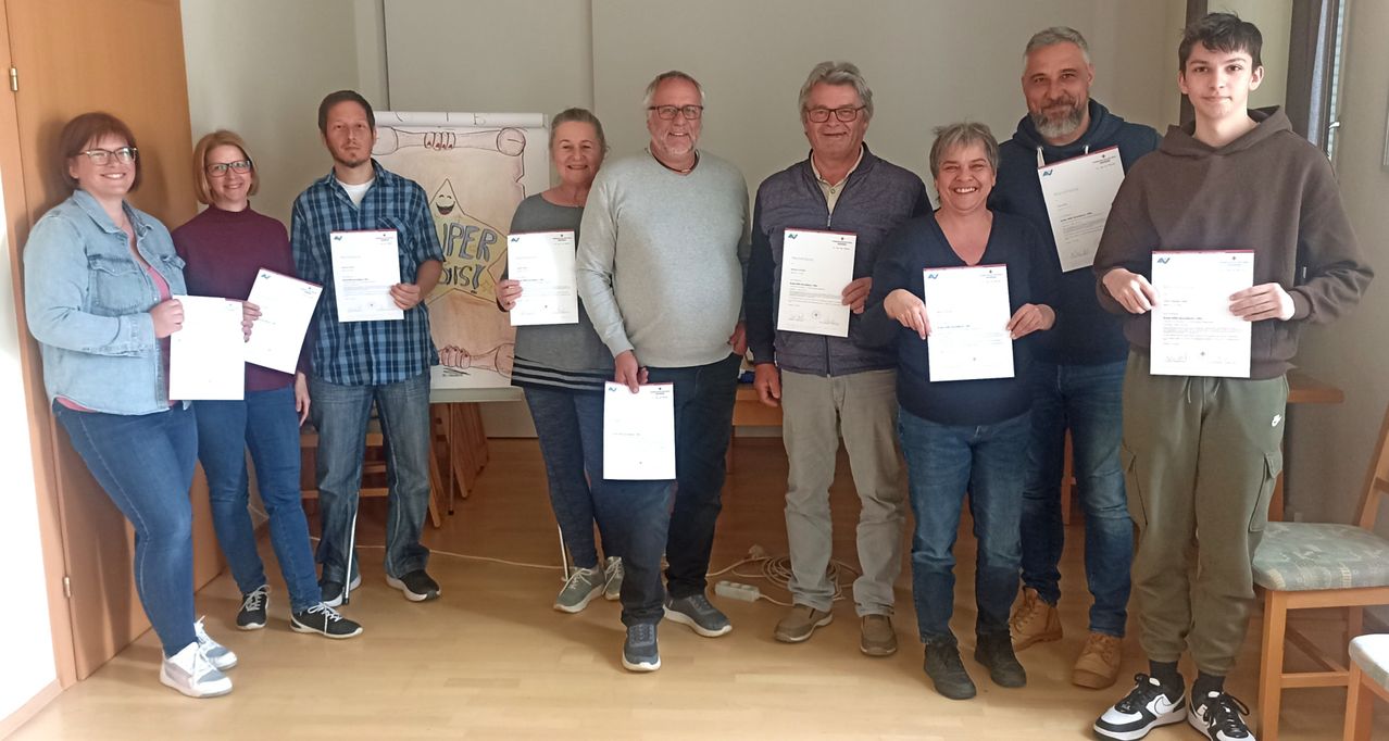 Sechs Erwachsene, sowohl Männer als auch Frauen, stehen mit Urkunden in der Hand, lächelnd und posieren für ein Foto. Dahinter befindet sich ein Plakat mit Text.