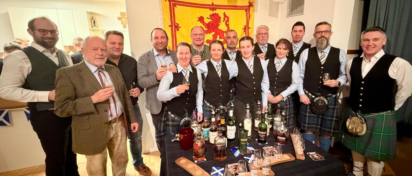 Gruppe von Menschen posiert vor einem Tisch mit Likörflaschen und Gläsern. Einige tragen Kilts. Schottische Flaggen sind im Hintergrund.
