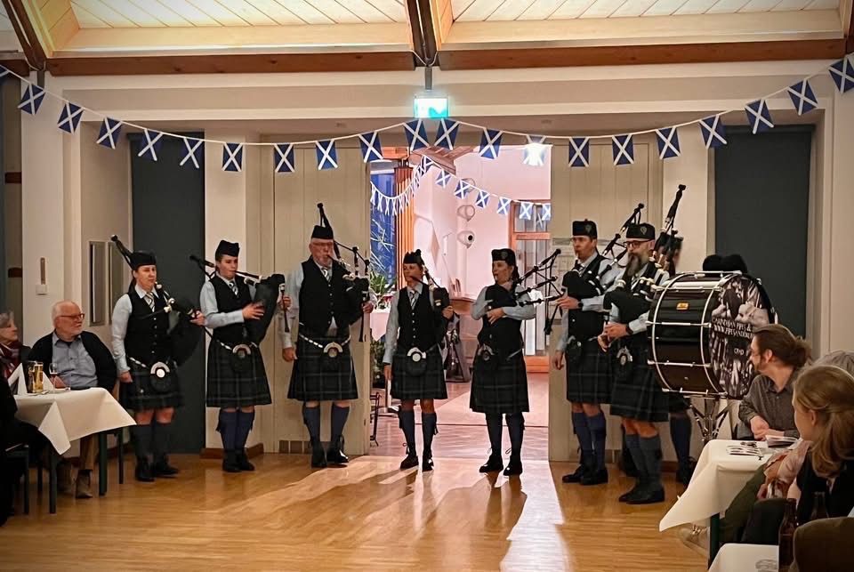 Eine Gruppe von Dudelsackspielern, die in Kilts und schwarze Stiefel gekleidet sind, tritt auf einer Bühne auf, vor ihnen steht ein Schlagzeug. Die Bühne hat Holzböden und einen Tisch mit einer weißen Tischdecke.