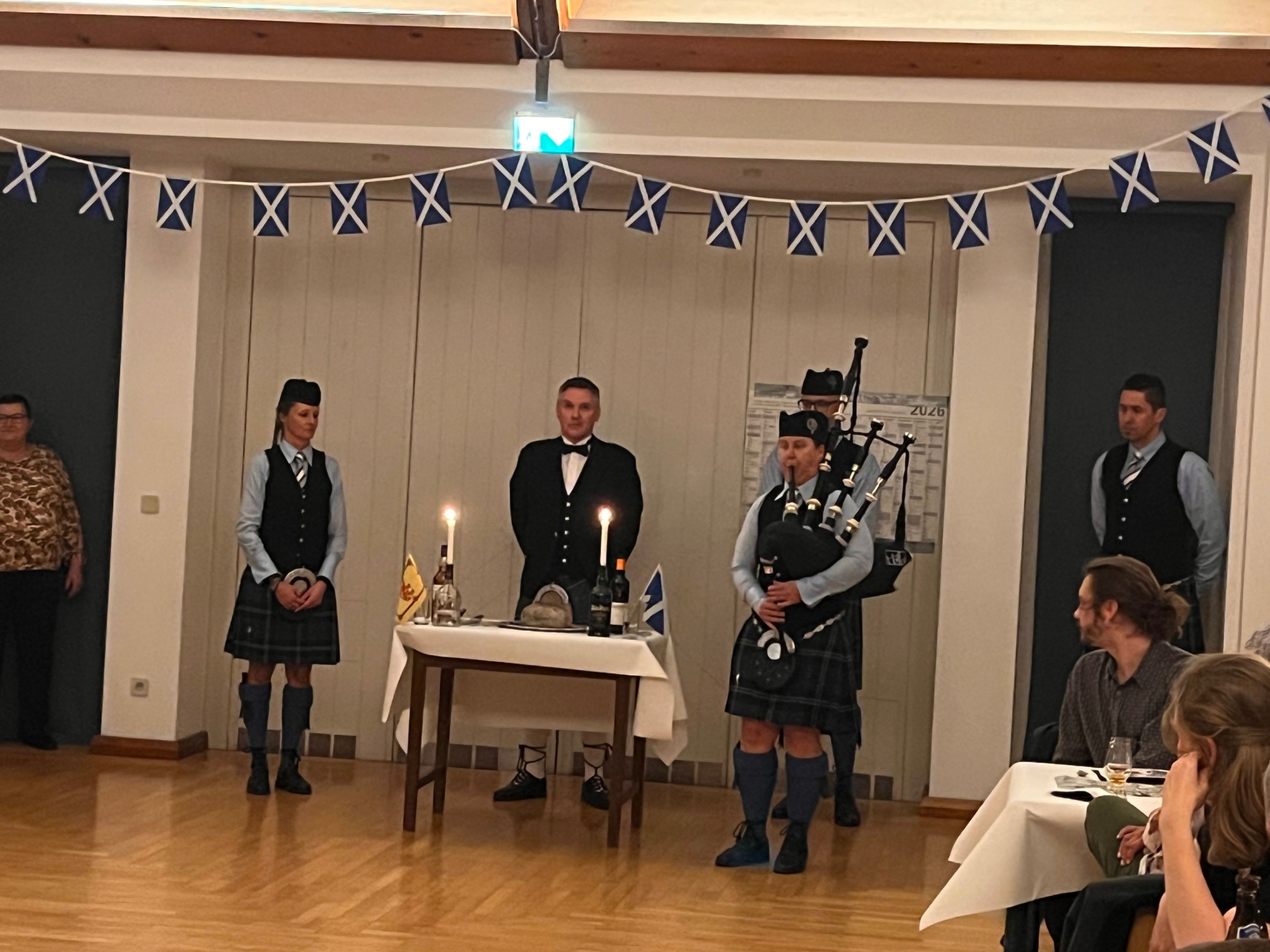 In einem Raum mit schottischen Flaggen spielt ein Mann Dudelsack, während ein Mann in formeller Kleidung an einem Tisch mit Kerzen steht. Eine weitere Person in einem Kilt steht in der Nähe.