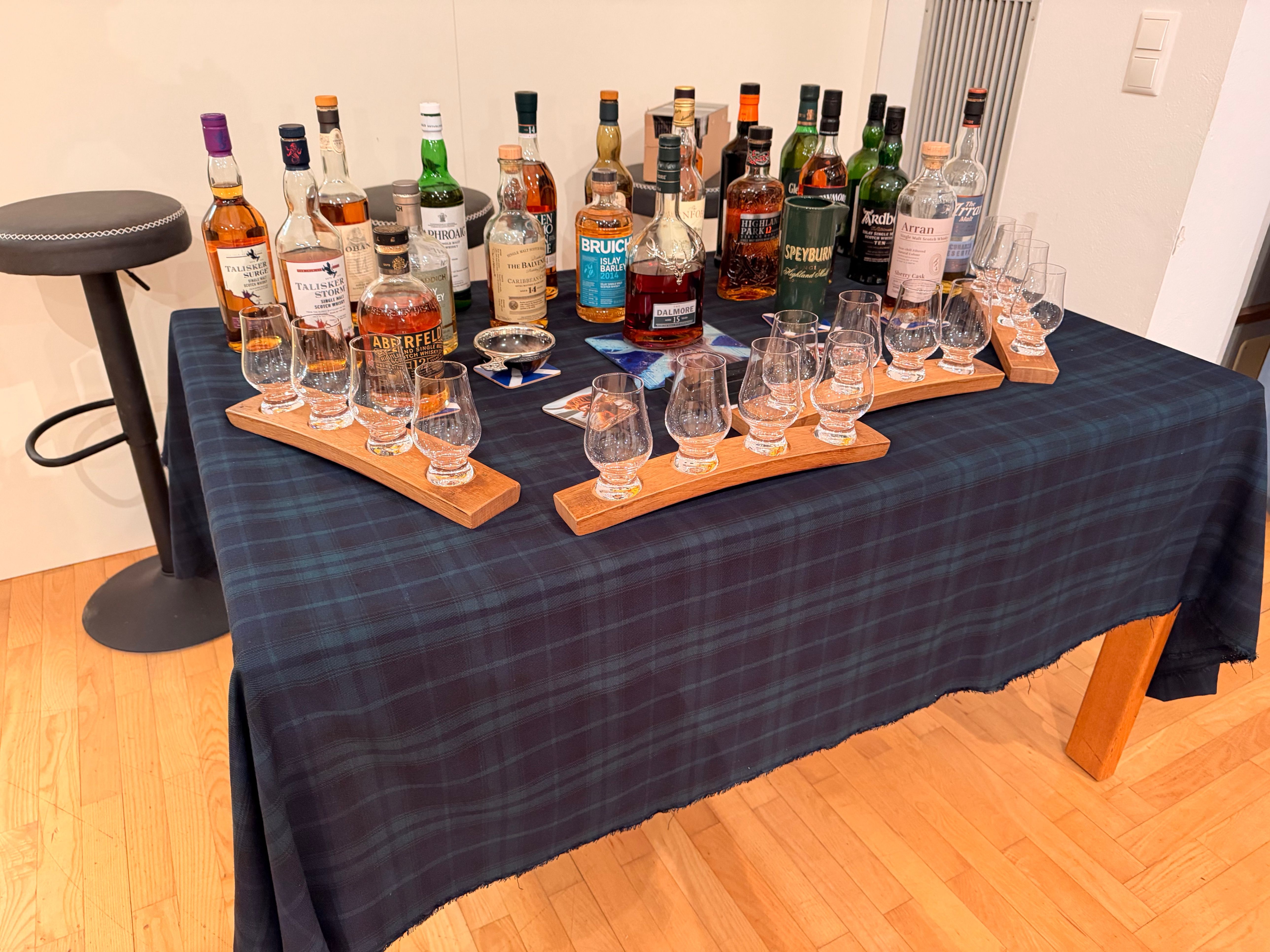 Ein Tisch mit einer karierten Tischdecke zeigt verschiedene Whiskyflaschen, einige mit Gläsern auf Holzbrettern. Die Umgebung ist drinnen mit Holzböden und weißen Wänden.