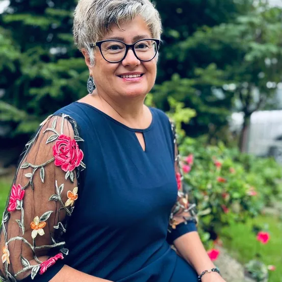 Eine Frau mit kurzen grauen Haaren, Brille, blauem Blumenkleid und Ohrringen sitzt in einem Garten.