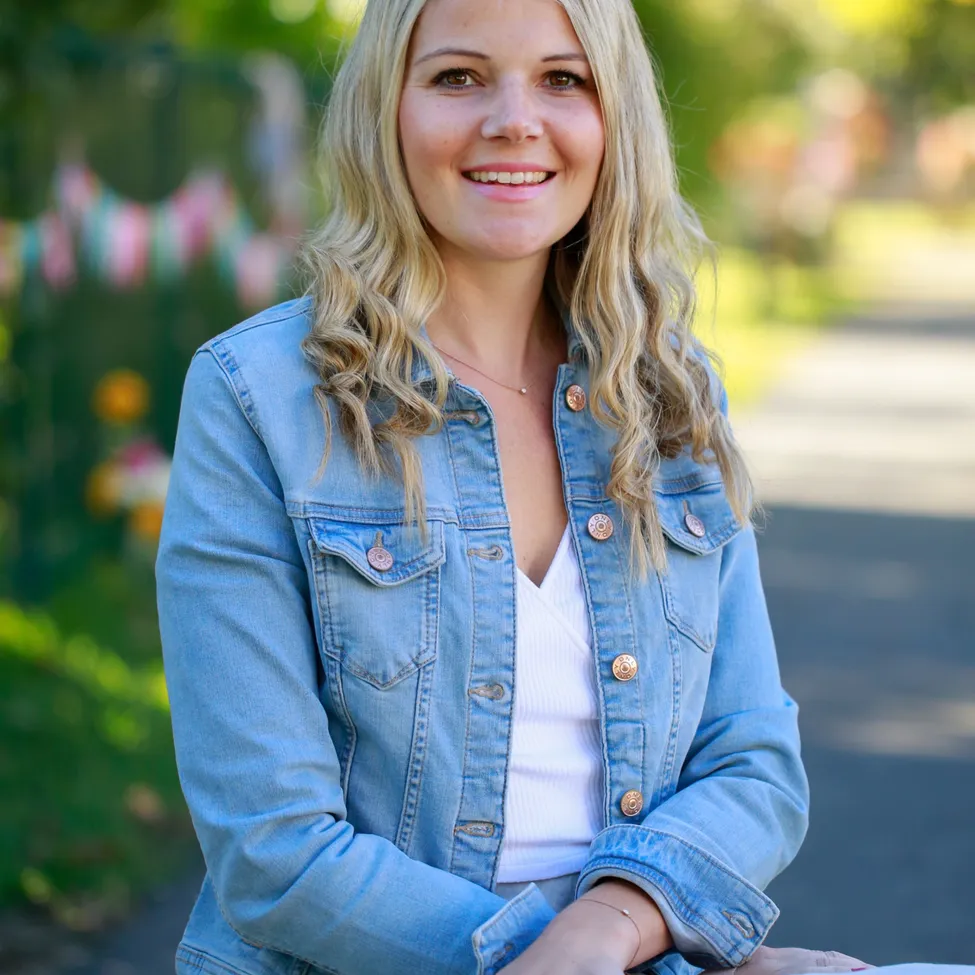 Eine Frau mit blonden Haaren, die ein weißes Shirt und eine Jeansjacke trägt, lächelt für ein Foto. Sie sitzt auf einem Weg mit Pflanzen und Blumen im Hintergrund.