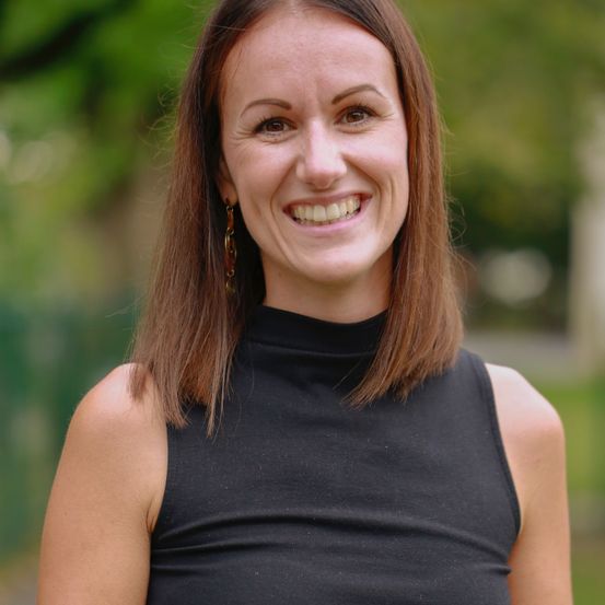 Eine lächelnde Frau mit braunen Haaren in einem ärmellosen schwarzen Top posiert für ein Foto im Freien.