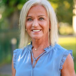 Eine Frau mit blonden Haaren, die ein blaues Oberteil und eine Perlenkette trägt, lächelt in einem Park.