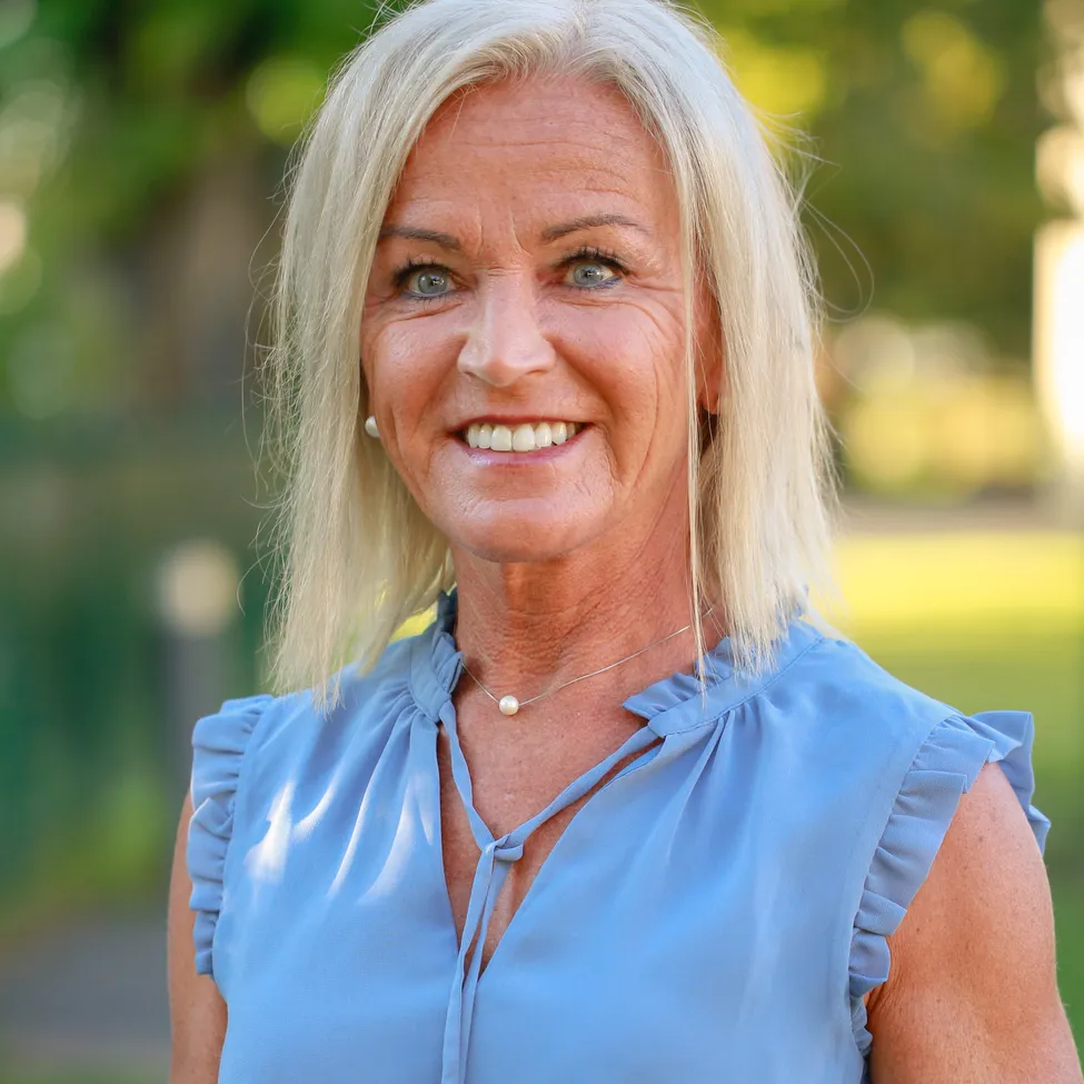 Eine Frau mit blonden Haaren, die ein blaues Oberteil und eine Perlenkette trägt, lächelt in einem Park.