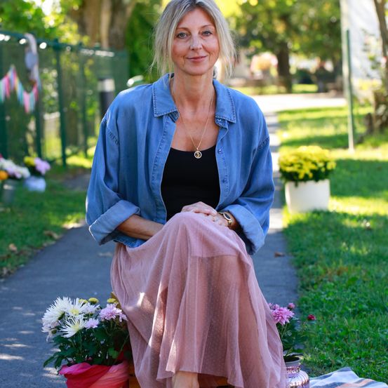 Eine Frau sitzt auf einem Bürgersteig, lächelt und trägt eine Jeansjacke und einen rosa Rock. Hinter ihr sind Blumen arrangiert, und der Hintergrund zeigt einen Rasenbereich mit Bäumen und einem Zaun.