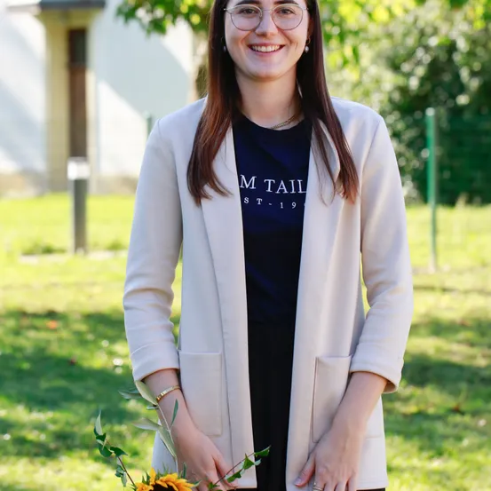 Eine lächelnde Frau mit Brille hält eine Sonnenblume. Sie trägt einen cremefarbenen Blazer und ein schwarzes T-Shirt. Im Hintergrund befinden sich ein Rasen und ein Gebäude.