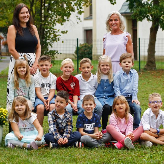 Eine Gruppe von Kindern und zwei Frauen, die draußen auf einem Rasenfeld posieren, für ein Foto vor einem weißen Gebäude und Bäumen im Hintergrund.