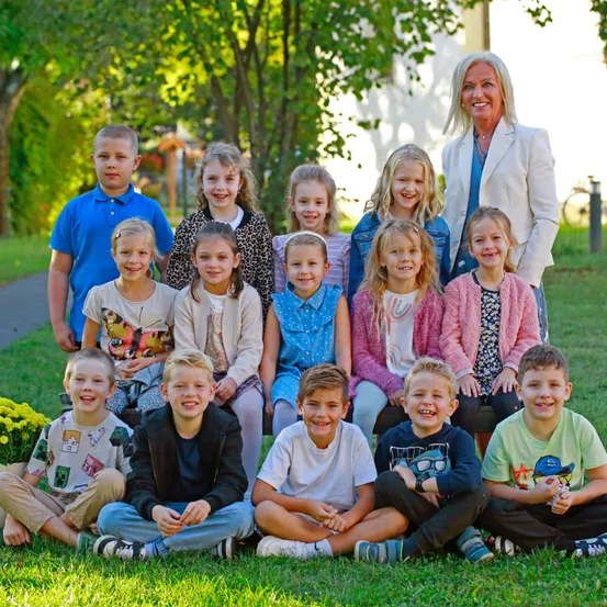 Eine Gruppe von Kindern und einer erwachsenen Frau posiert für ein Foto an einem sonnigen Tag. Die Kinder sitzen auf dem Rasen, während die Frau hinter ihnen steht.