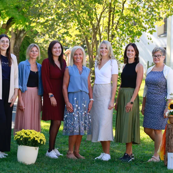 Eine Gruppe von Frauen in Kleidern und mit Brillen steht auf Gras mit einer weißen Vase und gelben Blumen, umgeben von Bäumen und einem Gebäude im Hintergrund.