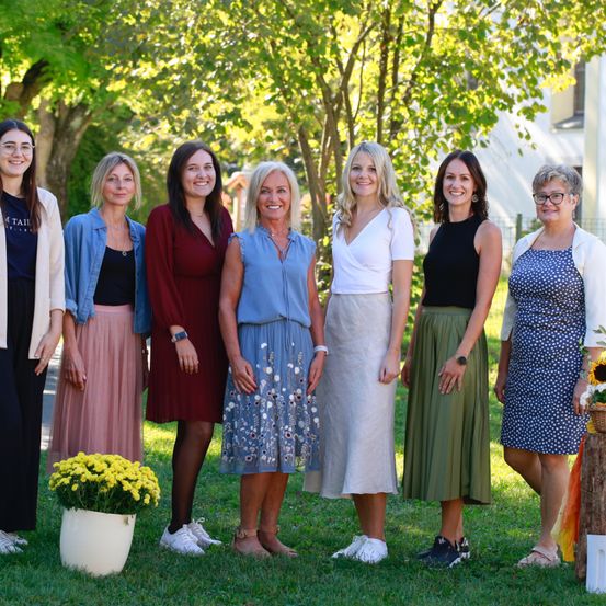 Eine Gruppe von Frauen in Kleidern und mit Brillen steht auf Gras mit einer weißen Vase und gelben Blumen, umgeben von Bäumen und einem Gebäude im Hintergrund.