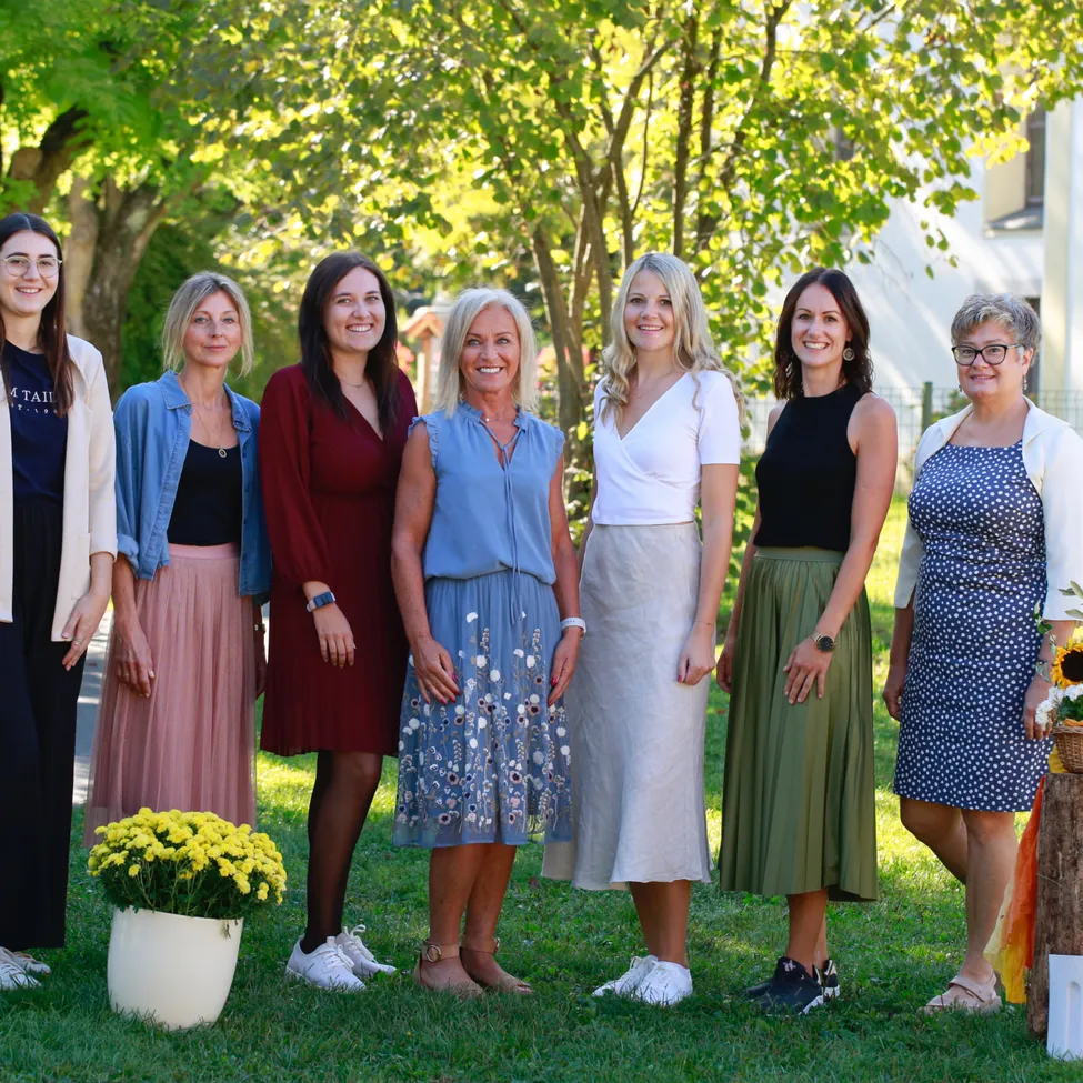 Eine Gruppe von Frauen in Kleidern und mit Brillen steht auf Gras mit einer weißen Vase und gelben Blumen, umgeben von Bäumen und einem Gebäude im Hintergrund.