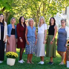 Eine Gruppe von Frauen in Kleidern und mit Brillen steht auf Gras mit einer weißen Vase und gelben Blumen, umgeben von Bäumen und einem Gebäude im Hintergrund.