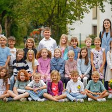 Eine Gruppe von Kindern in verschiedenen Kleidungsstücken posiert für ein Foto in einem Park mit einem Gebäude im Hintergrund.