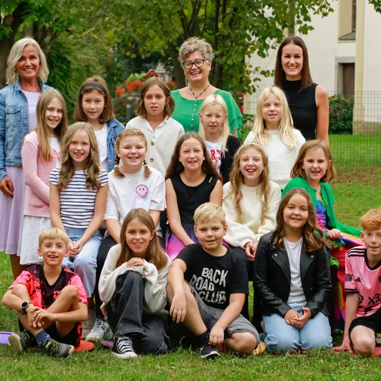 Eine Gruppe von Kindern und Erwachsenen steht in einem Rasenbereich mit Bäumen im Hintergrund. Sie lächeln und posieren für ein Foto.