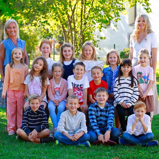 Ein Gruppenfoto einer Klasse von Kindern und zwei Lehrerinnen auf einem Rasenfeld mit Bäumen im Hintergrund.