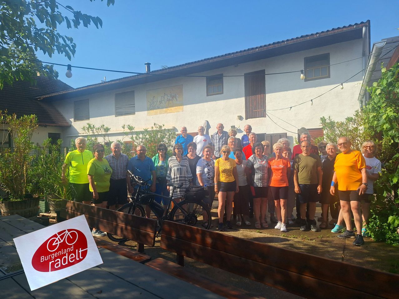Eine Gruppe von Menschen steht vor einem Gebäude mit Fahrrädern und lächelt für ein Foto. Links ist ein Schild mit der Aufschrift 'Ergenland Radelt' zu sehen.