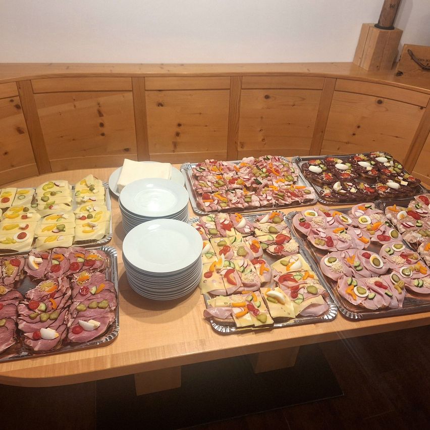 Ein Holztisch zeigt verschiedene Platten mit Sandwiches, einschließlich Schinken, Käse und Essiggurken. In der Nähe sind Teller und Schüsseln gestapelt. Im Hintergrund befinden sich Holzschränke.