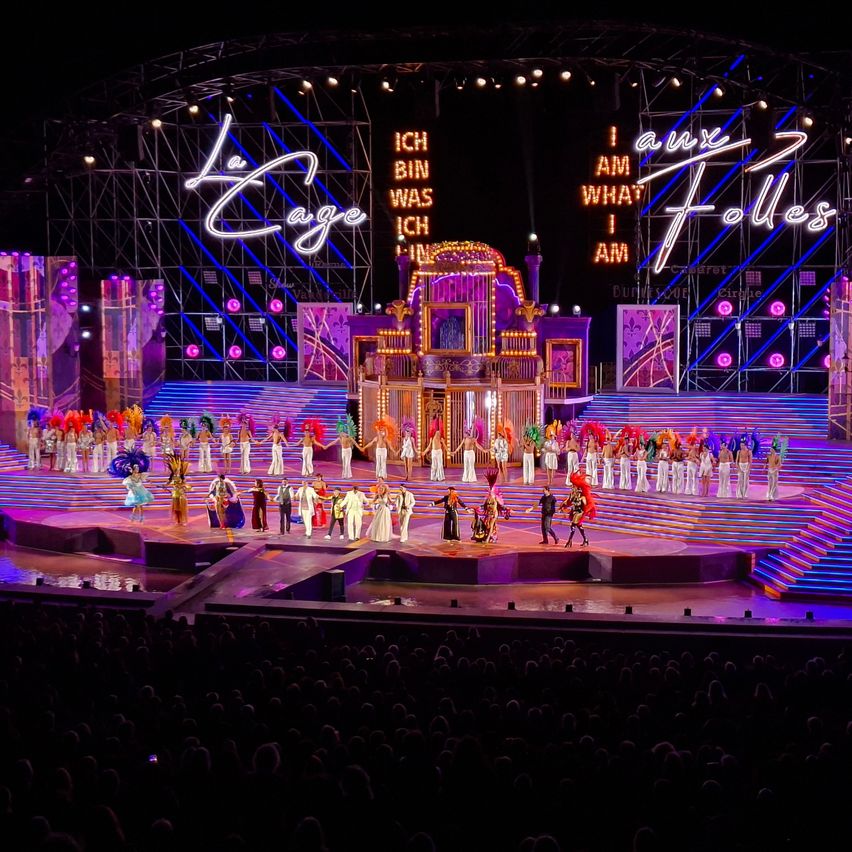 A stage performance with a large audience, featuring performers in colorful costumes and a vibrant stage setup with neon lights and a prominent sign reading 'La Cage aux Folles'.