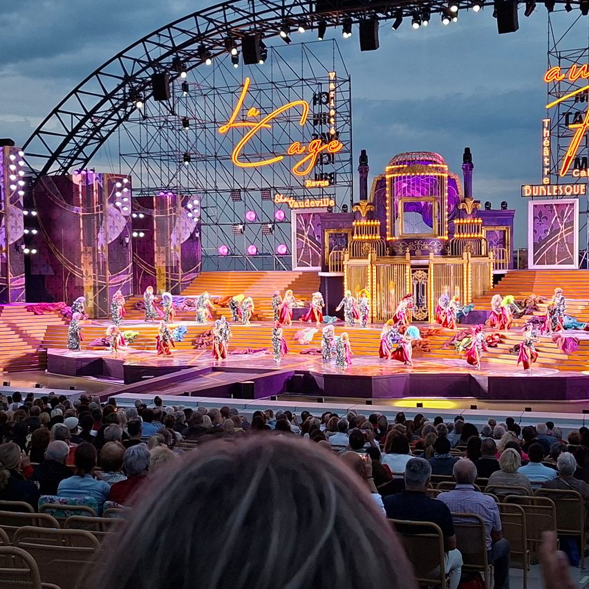 A vibrant stage performance under a large arch with 'La Cage' written in yellow. Dancers in colorful costumes are performing on a multi-level stage with an audience seated below.