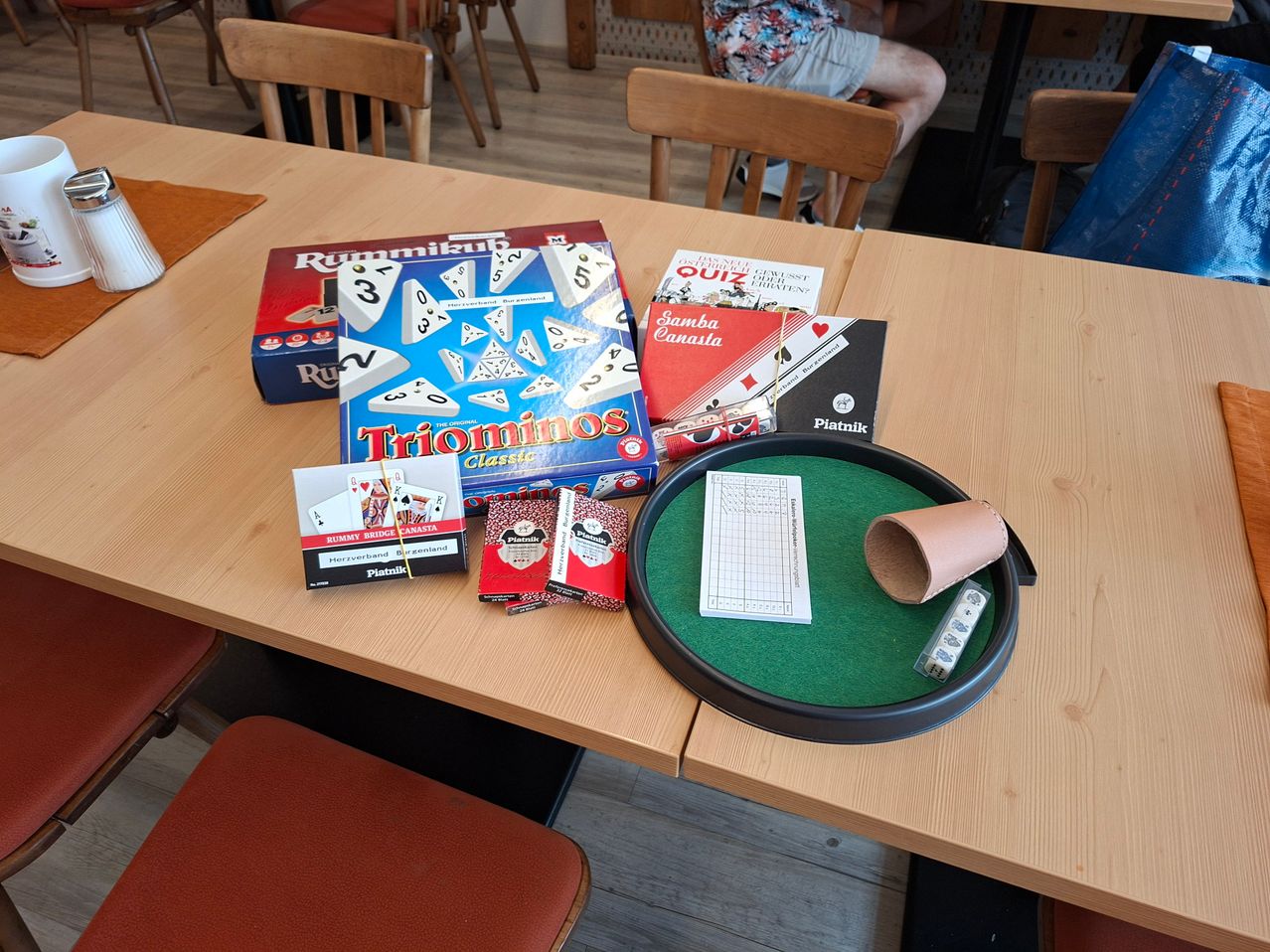 A wooden table displays various board games and accessories, including Rummikub, Triominos, Canasta cards, and a green felt game board.