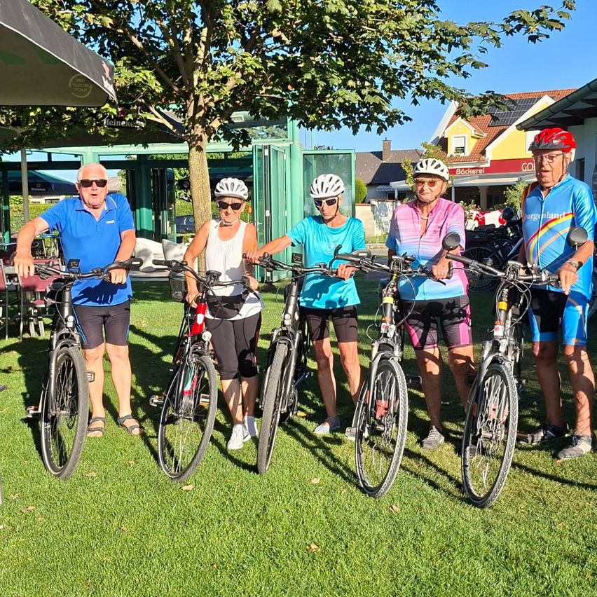 Fünf Radfahrer, alle mit Helmen, stehen auf einer Rasenfläche. Sie lächeln und scheinen für ein Foto zu posieren. Hinter ihnen befinden sich Gebäude und Bäume.