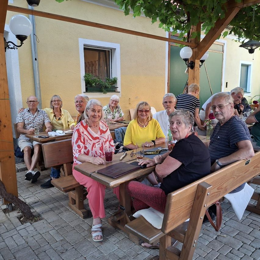 Eine Gruppe älterer Menschen sitzt an einem Tisch vor einem Restaurant. Sie lächeln und scheinen ihre Mahlzeit zu genießen. Der Tisch ist mit Tellern, Gläsern und Servietten gedeckt.