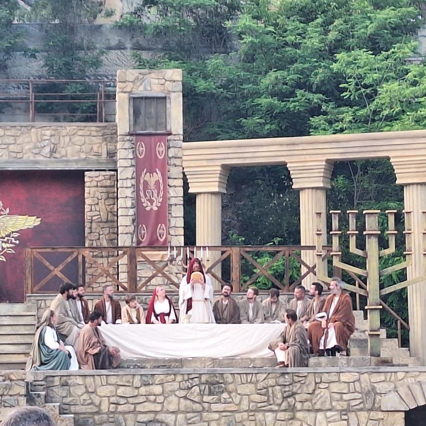 A group of people sit around a table on a stage, dressed in ancient costumes, with a backdrop of stone and greenery. A central figure holds a bread.