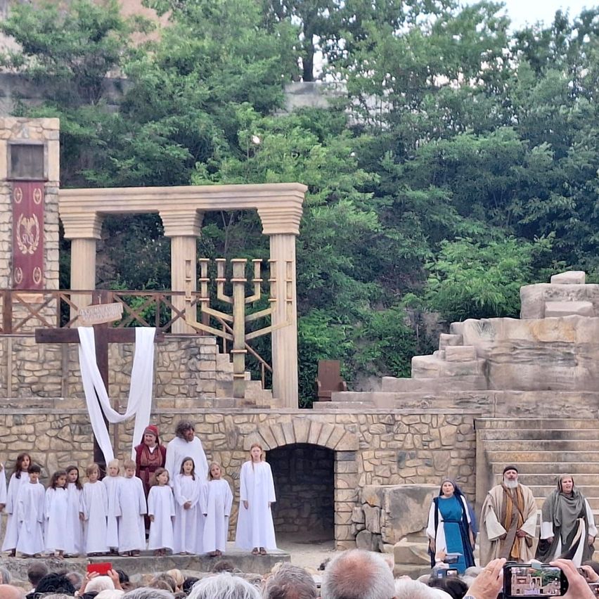 A group of performers on a stone stage with a backdrop of trees, featuring a woman in a blue dress and two men in robes. A choir of children in white robes stand before them.