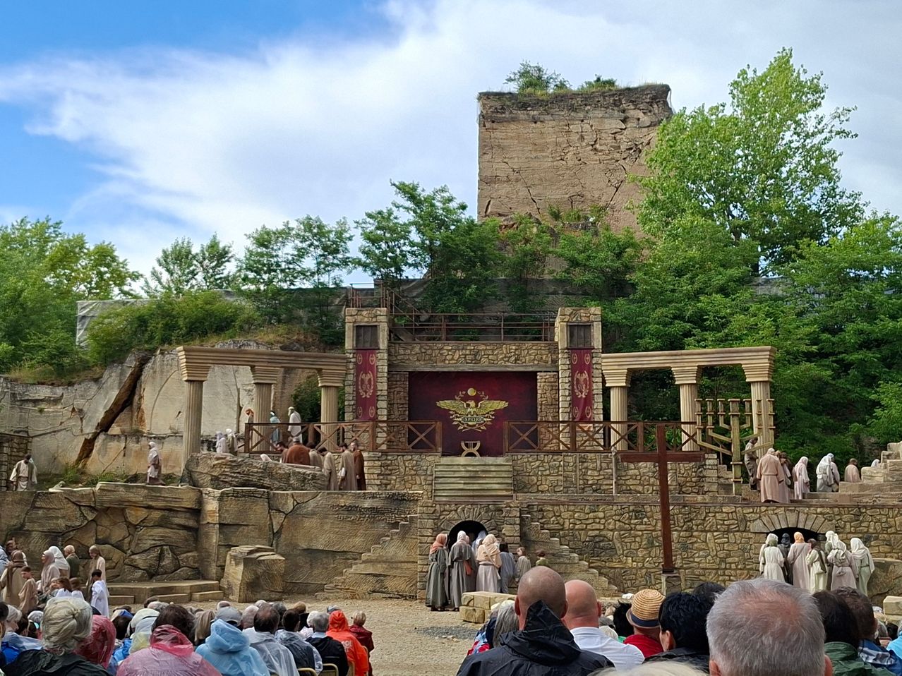 A historical reenactment event with an audience seated in front of a stone structure. Actors dressed in period costumes perform on a stage with a red backdrop. Trees and a ruined tower are visible in the background.