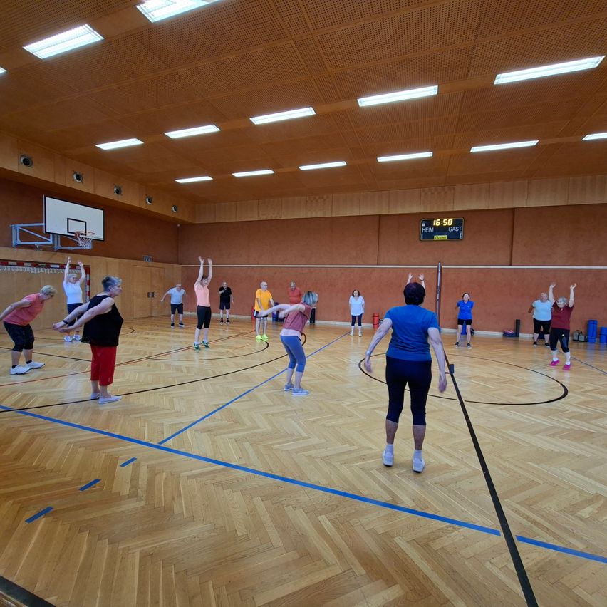 Eine Gruppe von Menschen übt in einer Turnhalle. Sie machen eine Hula-Hoop-Routine. Der Boden ist aus Holz. Ein Basketballkorb ist an der Wand montiert. Eine Anzeigetafel ist an der Wand montiert.