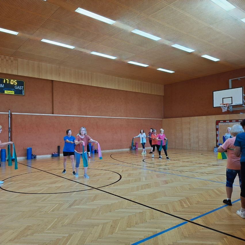 Eine Gruppe älterer Frauen trainiert in einem Innengym. Sie halten Bänder und scheinen Anweisungen zu befolgen. Das Fitnessstudio verfügt über ein Basketballfeld und eine Anzeigetafel.