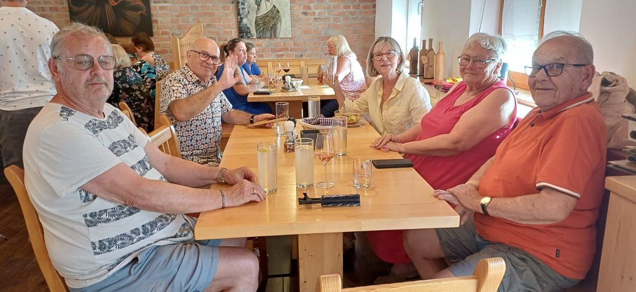Eine Gruppe älterer Erwachsener sitzt an einem Holztisch, lächelt und hebt ein Glas Wein. Sie sind in einem Restaurant mit Ziegelwänden und einem Gemälde an der Wand.