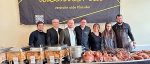 Eine Gruppe von Menschen steht hinter einem mit Fleisch bedeckten Tisch. Sie tragen Schürzen und Brillen. Ein Banner mit dem Text ZWOA Weinvienta ist im Hintergrund.