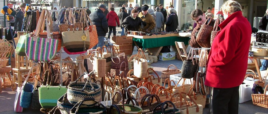Ein belebter Straßenmarkt mit zahlreichen ausgestellten geflochtenen Körben, einkaufenden und stöbernden Menschen, einem Verkäufertisch mit Waren und Gebäuden im Hintergrund.