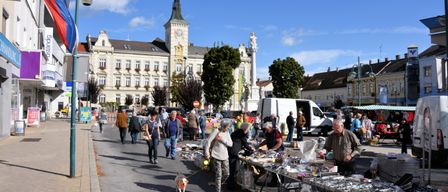 Freiluftmarkt mit verschiedenen Ständen und gehenden Menschen an einem sonnigen Tag, mit einem großen Gebäude und einem Uhrturm im Hintergrund.