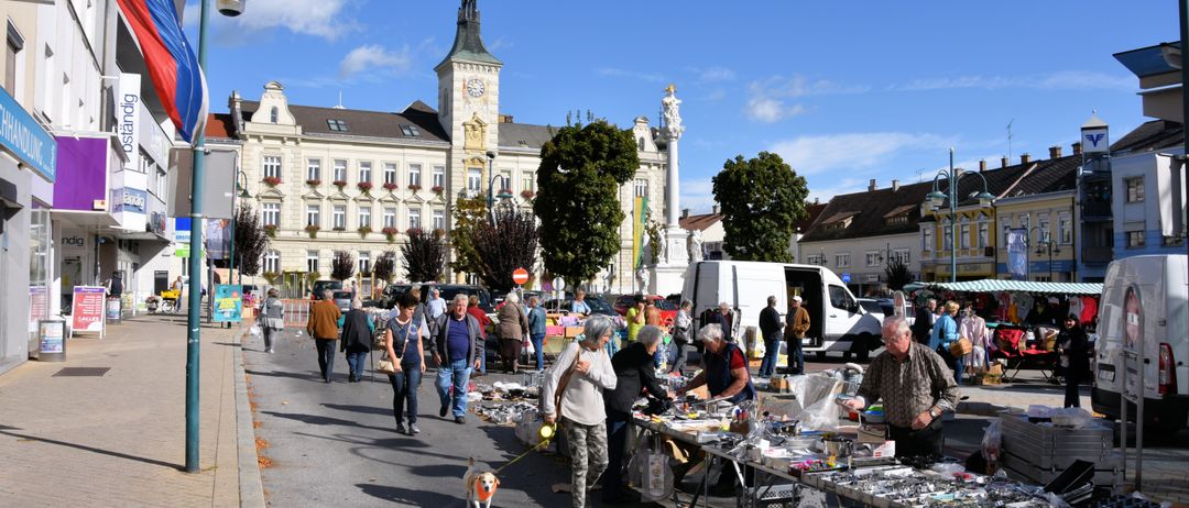Freiluftmarkt mit verschiedenen Ständen und gehenden Menschen an einem sonnigen Tag, mit einem großen Gebäude und einem Uhrturm im Hintergrund.