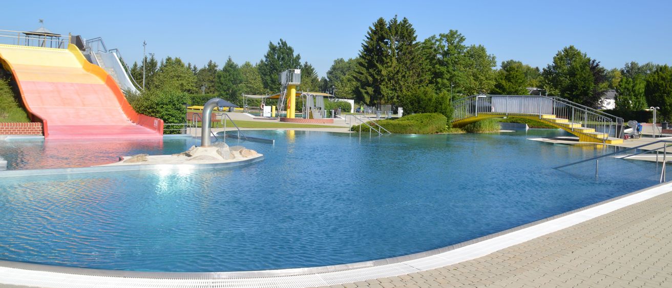 Ein großer Außenpool mit Wasserrutsche, umgeben von Grün und Bäumen, unter einem klaren blauen Himmel.