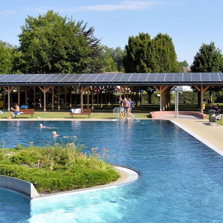 Ein Freibad mit Solarpanelen darüber, umgeben von Grünflächen und einem Holzpavillon. Einige Menschen schwimmen und andere entspannen sich auf der Wiese.