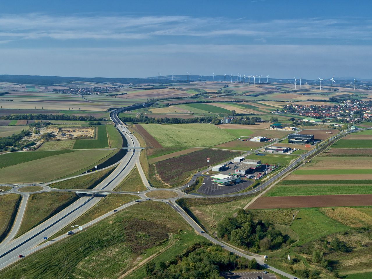 Luftaufnahme einer Autobahnkreuzung mit Autos auf der Straße, umgeben von landwirtschaftlichen Feldern, Bäumen und einer Tankstelle. In der Ferne befinden sich Windräder am Horizont.