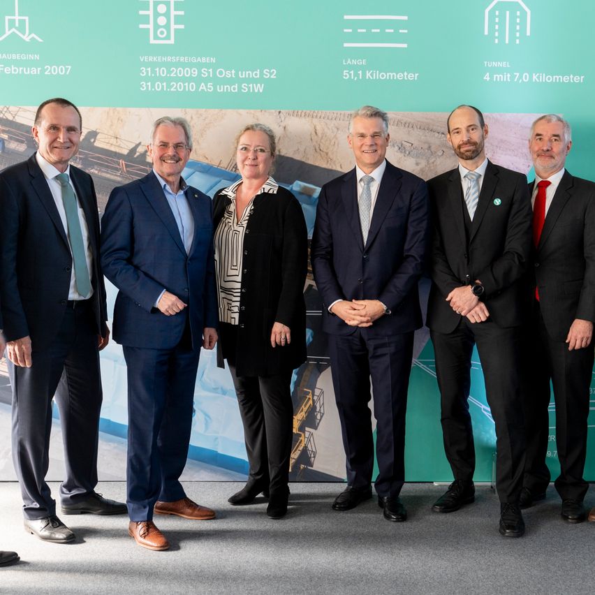 Fünf Männer und eine Frau in Anzug stehen nebeneinander vor einem Banner mit Grafiken und Text.