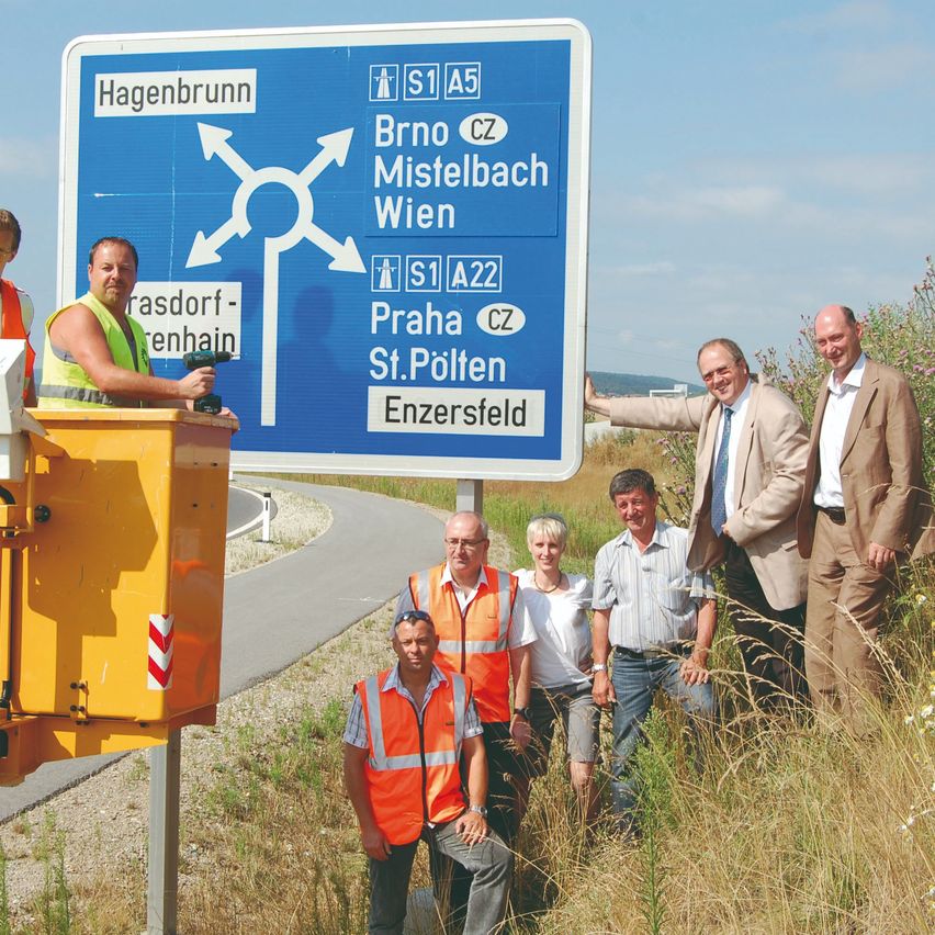 Eine Gruppe von Männern und Frauen, einige in Warnwesten, posiert neben einem Straßenschild mit einer gelben Basis. Das Schild zeigt verschiedene Routen und Ziele, darunter Hagenbrunn, Mistelbach, Wien, Prag, St. Pölten und Enzersdorf.
