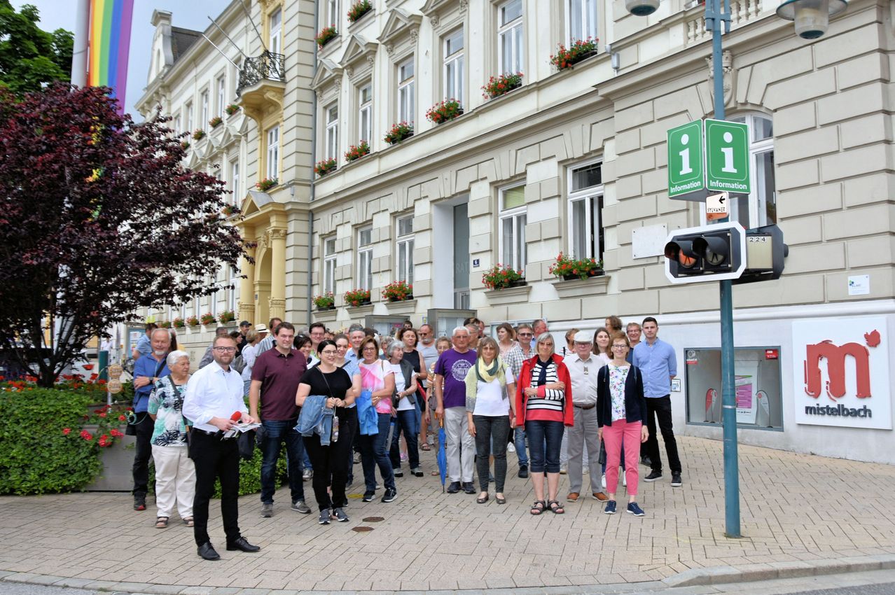 Eine Gruppe von Menschen steht vor einem weißen Gebäude mit mehreren Fenstern und Blumen auf den Balkonen. Die Straße hat eine Ampel und ein Informationsschild.